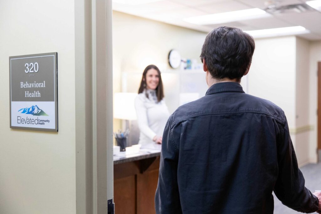 Person entering a behavioral health office at Elevated Community Health, showcasing a welcoming reception area with a staff member smiling behind the desk.