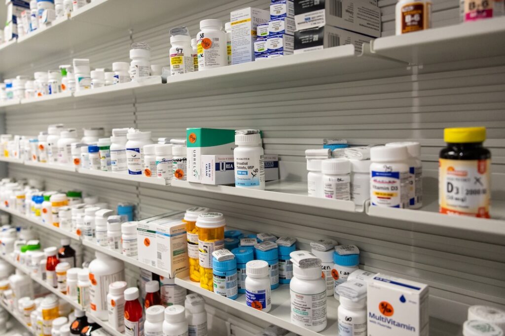 Shelves filled with various medication bottles, including prescription drugs and vitamins, organized by type and labeled for easy identification in a pharmacy setting.