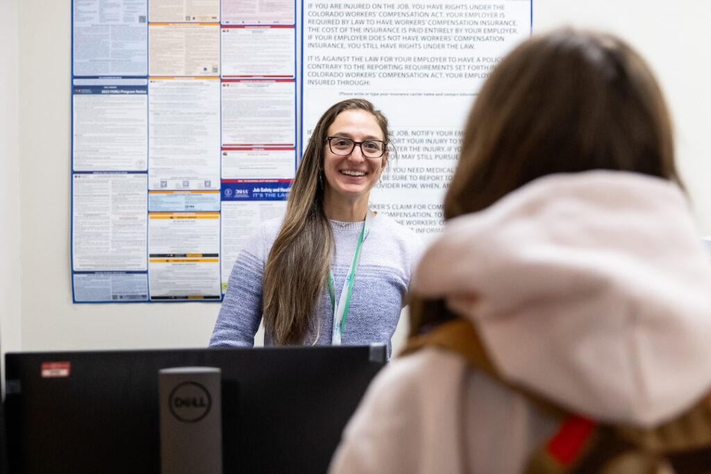 Smiling woman at a reception desk providing information, with workplace safety posters displayed on the wall behind her. A visitor in a hooded sweatshirt is facing her, engaged in conversation.