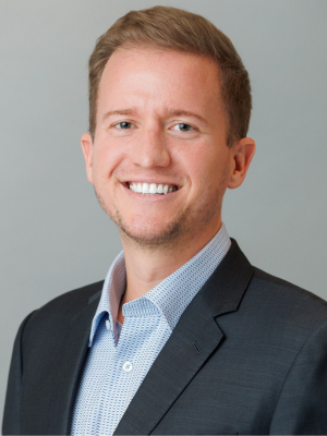 Professional headshot of a smiling young man wearing a black suit jacket over a light blue patterned shirt, set against a neutral gray background.