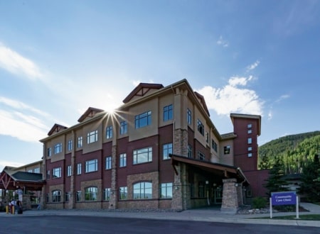 Modern healthcare facility in a scenic mountain setting, featuring large windows and stone accents, with ample parking and sustainable architecture.