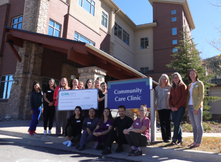 Group of healthcare workers and staff standing outside the Community Care Clinic, holding a donation check, with the clinic building prominently displayed in the background.
