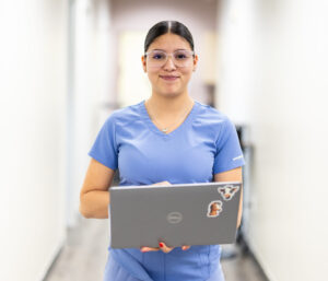 Young woman in blue scrubs holding a laptop in a brightly lit hallway, representing healthcare professionalism and technology use in medical settings.