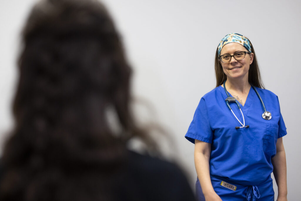 Healthcare professional in blue scrubs smiles while interacting with a patient. The setting suggests a clinical environment, emphasizing patient care and communication.