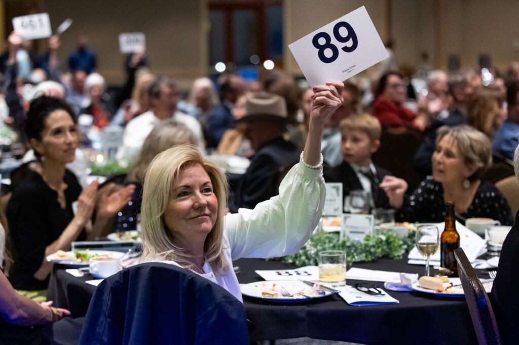 Woman holding a bid card with the number 89 at a lively fundraising event, surrounded by an engaged audience at elegantly set tables, showcasing a social atmosphere.