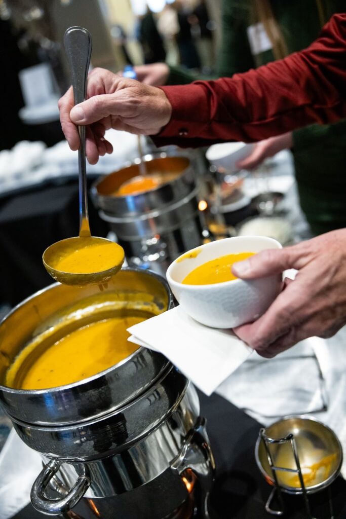 A person serving creamy orange soup from a stainless steel pot into a white bowl at a buffet, showcasing a warm dining atmosphere.
