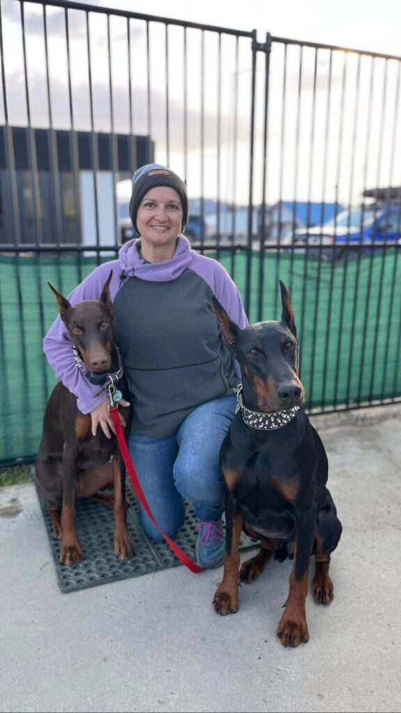 Woman sitting with two Doberman dogs in a fenced outdoor area, wearing a purple and gray sweater with a beanie, showcasing a friendly bond with the pets.
