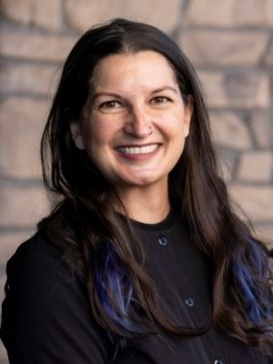 Portrait of a woman with long dark hair featuring blue highlights, smiling warmly against a stone wall background. She is wearing a black shirt and has a friendly demeanor.