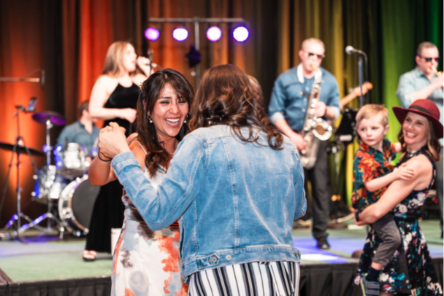 A vibrant dance scene at a lively event featuring two women joyfully interacting on the dance floor, with a live band performing in the background. The atmosphere is festive, highlighted by colorful stage lighting, while a child is seen being held by a woman nearby.